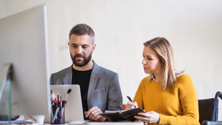 Office worker in wheelchair writing on notepad and working on computer with coworker